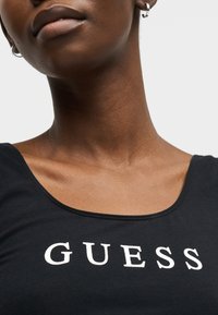 Κοντινό πλάνο ενός ατόμου που φορά ασημένια κρεμαστά σκουλαρίκια και μαύρο τοπ με το λευκό λογότυπο "GUESS" στο στήθος.