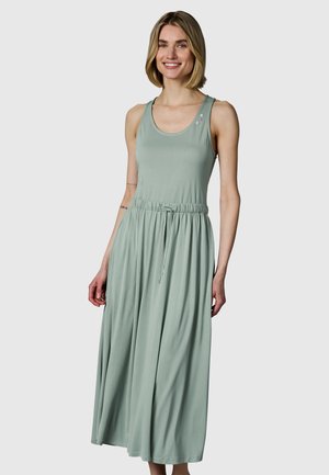 Lächelnde Frau mit kurzen blonden Haaren, die ein ärmelloses, hellgrünes Maxikleid mit Kordelzug in der Taille und Knöpfen am Ausschnitt trägt.