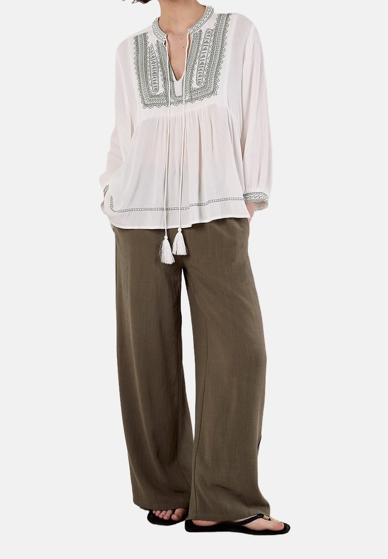 Femme portant un chemisier blanc brodé avec des pompons, un pantalon ample couleur olive et des tongs noires, debout les mains dans les poches.