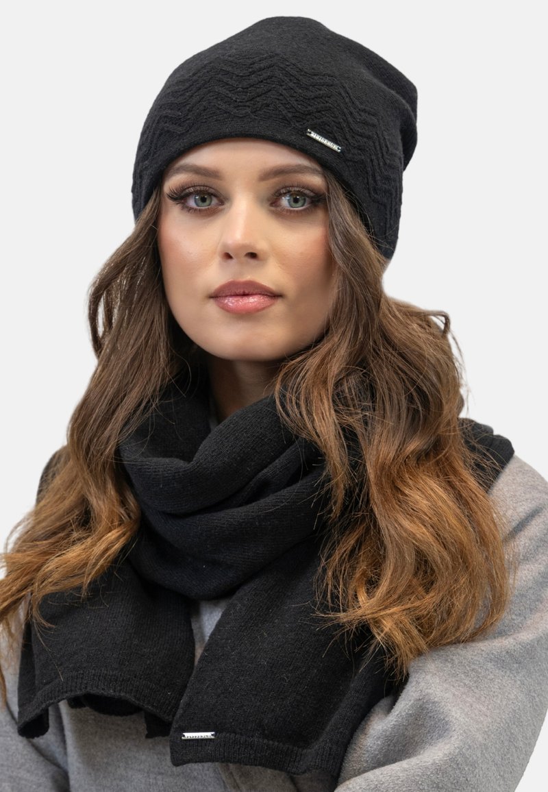 Cappello a maglia nera con motivo a onde e un logo argentato, abbinato a una sciarpa nera, entrambi in tessuto morbido, indossati sopra un maglione grigio chiaro.