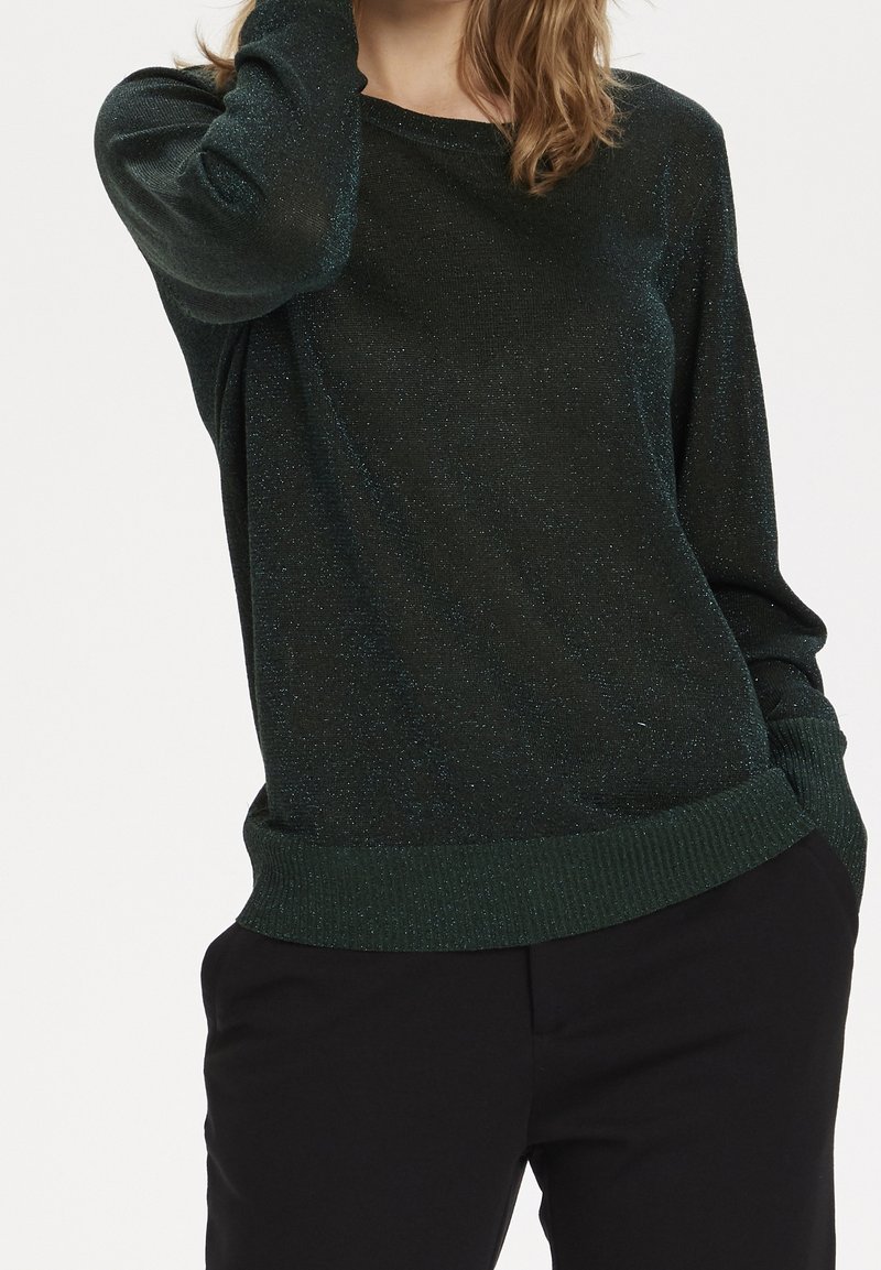 Femme portant un pull vert foncé pailleté avec des poignets et un ourlet côtelés, associé à un pantalon noir, posant avec une main près de sa tête.
