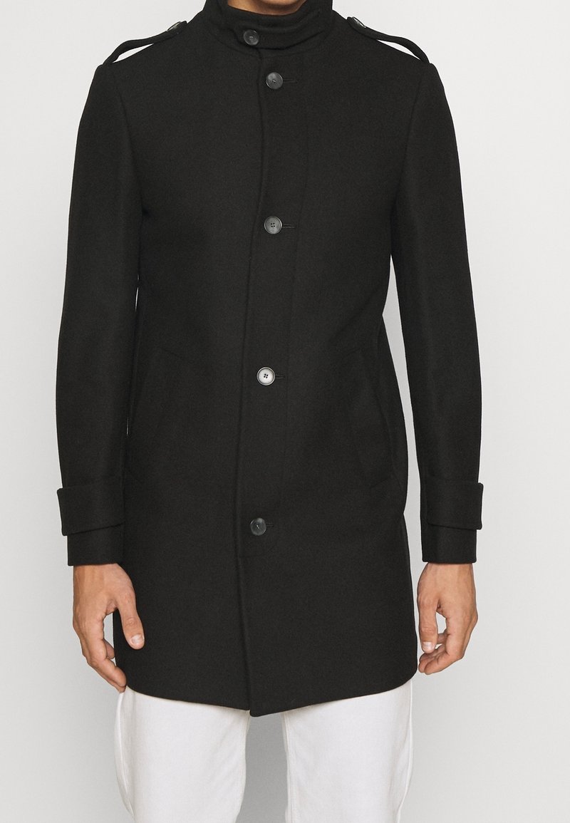Zwarte wollen jas met een hoge kraag, knopenpat, klepzakken en schouderepauletten. Soepele textuur, getailleerde pasvorm.