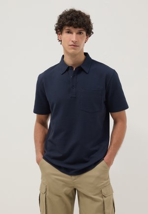 Joven con cabello rizado, de pie con las manos en los bolsillos, vestido con una camisa polo azul marino de manga corta y pantalones cargo beige sobre un fondo liso.