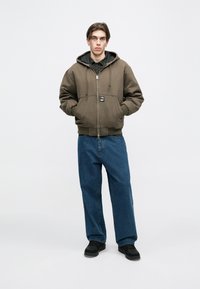 Maslinasto zelena jakna s kapuljačom i prednjim patentnim zatvaračem, s dva bočna džepa, nošena preko crne košulje na kockice i plavih hlača širokih nogavica, uparena s crnim cipelama.