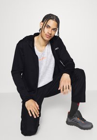 Felpa nera con zip sopra una maglietta grigia in rete, pantaloni neri e scarpe grigie con dettagli arancioni. Capelli intrecciati acconciati vicino alla testa.