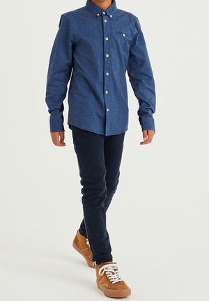 Niebieska, dżinsowa koszula z długim rękawem na guziki, zestawiona z ciemnymi spodniami i brązowymi trampkami. Koszula posiada kieszeń na piersi i białe guziki.