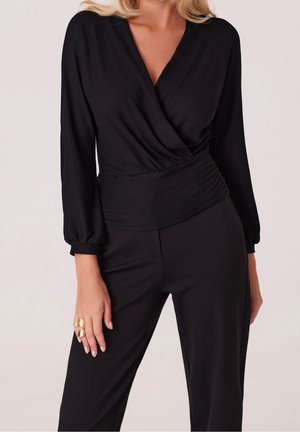 Kvinde iført en sort langærmet omslagstop med rynket talje kombineret med sorte bukser, der viser en guldring på hendes venstre hånd.