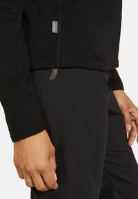 Hænder ned langs siderne iført sort langærmet top med "Icebreaker"-mærke og sorte bukser med lynlåslomme-detalje.