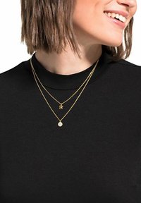 Guldpläterade halsband med en stjärnformad berlock och en rund, glittrig charm mot en svart topp. Texturerade metallkedjor.