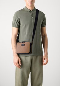 Brązowa, teksturowana torba na ramię o prostokątnym kształcie, z czarnymi elementami i zamkiem błyskawicznym, noszona przez osobę w casualowym stroju.