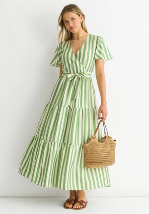 Woman wearing a green and white vertically striped maxi dress with short sleeves and a waist tie, holding a woven straw handbag and wearing sandals.