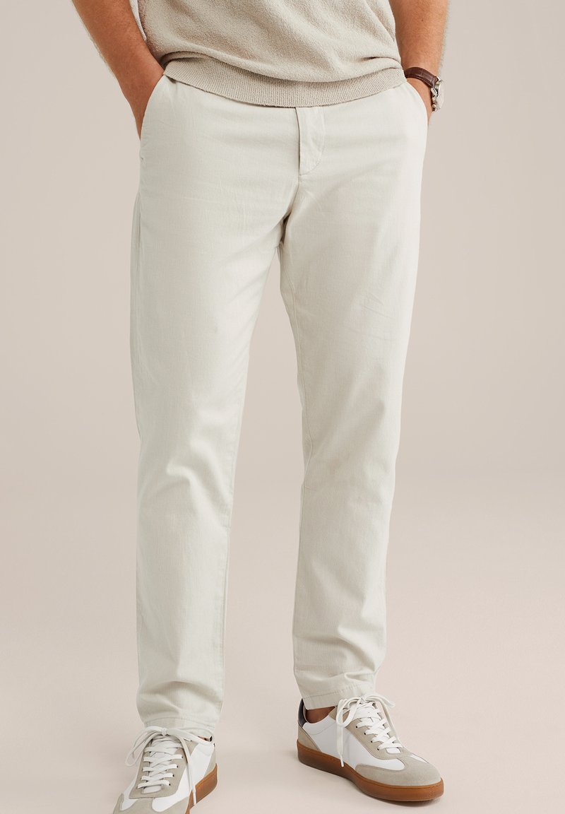 Pantaloni di cotone beige chiaro con vestibilità dritta, due tasche laterali e una texture liscia, abbinati a sneakers bianche e grigie.