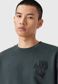 Zelená mikina s kulatým výstřihem, vyrobená z měkkého materiálu. Na levém prsu má výrazný černý grafický potisk. Minimalistický design.