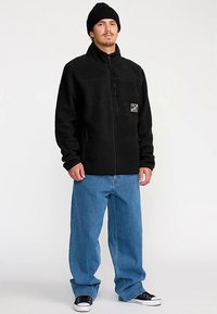 Zwarte fleecejas met hoge kraag, ritssluiting aan de voorkant en een borstzak. Gecombineerd met blauwe wijde jeans en zwarte sneakers. Gebreide zwarte muts.