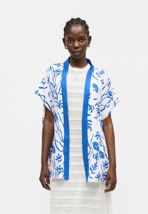 Punutud juustega naisel on seljas valge põlvpikkune kleit ja lahtine valge kimonoga, sinise lillelise mustri ja sinise äärega, seisab käed lõdvestunult.