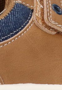 Zapatilla deportiva de cuero marrón con acentos de mezclilla, que presenta costuras y ojales en blanco. La textura es suave con un acabado delicado.