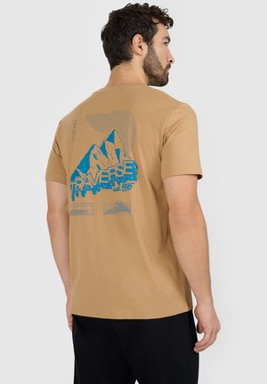 Mężczyzna w beżowej koszulce z niebieską grafiką góry i napisem „TRAVERSE est. 1995” na plecach, zwrócony bokiem na szarym tle.