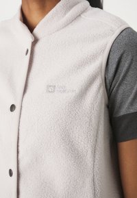 Persona que lleva un chaleco de forro polar beige claro con botones de presión sobre una camisa gris de manga corta, mostrando el logo de Jack Wolfskin en el pecho.