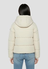 Beige dunjakke med hætte, quiltet design og elastiske ærmekanter, kombineret med lyseblå jeans.