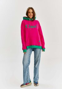 Różowa bluza z kapturem, z zielonymi akcentami, z napisem "Ciao!" w pogrubionych literach, kangurzą kieszenią i sznurkiem w kapturze, w połączeniu z jasnoniebieskimi dżinsami.