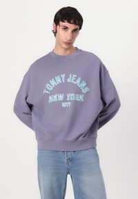 Purple cotton sweatshirt with "TOMMY JEANS NEW YORK CITY" embroidered in blue. Relaxed fit with ribbed cuffs and hem. Worn with light denim.