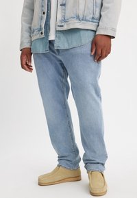 Vaqueros de denim azul claro con un corte relajado, combinados con una chaqueta de denim azul claro y zapatos tipo slip-on beige.
