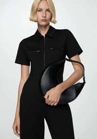 Sort halvmåneformet taske lavet af tekstureret læder, holdt af en person iført en kortærmet sort jumpsuit med lynlåslommer.