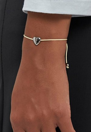 Adjustable bracelet featuring a silver heart charm on a thin, light-colored cord. The hand shown has a natural skin texture.