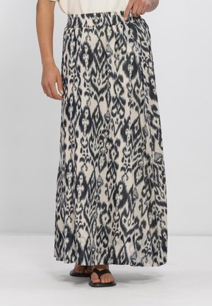 Vrouw die een zwart-witte maxi-rok met een elastische taille en zwarte sandalen draagt, staand tegen een effen achtergrond.