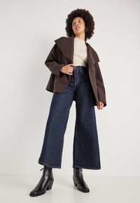 Cappotto oversize marrone con ampio colletto, maglioncino beige a coste, jeans a vita alta blu scuro a gamba larga e stivaletti neri con tacco.