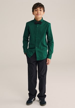 Groene gemuste blouse met een zwarte vlinderdas, gecombineerd met zwarte broeken en zwarte sneakers. Glad stof, aansluitend ontwerp.