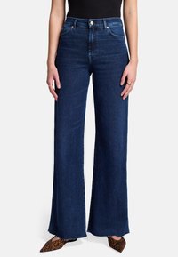 Jeans w wysokim stanie i szerokich nogawkach w ciemnoniebieskim kolorze z surowym wykończeniem, z przednimi kieszeniami i zapięciem na guzik. Połączone z butami w kropki z wężowym nadrukiem.