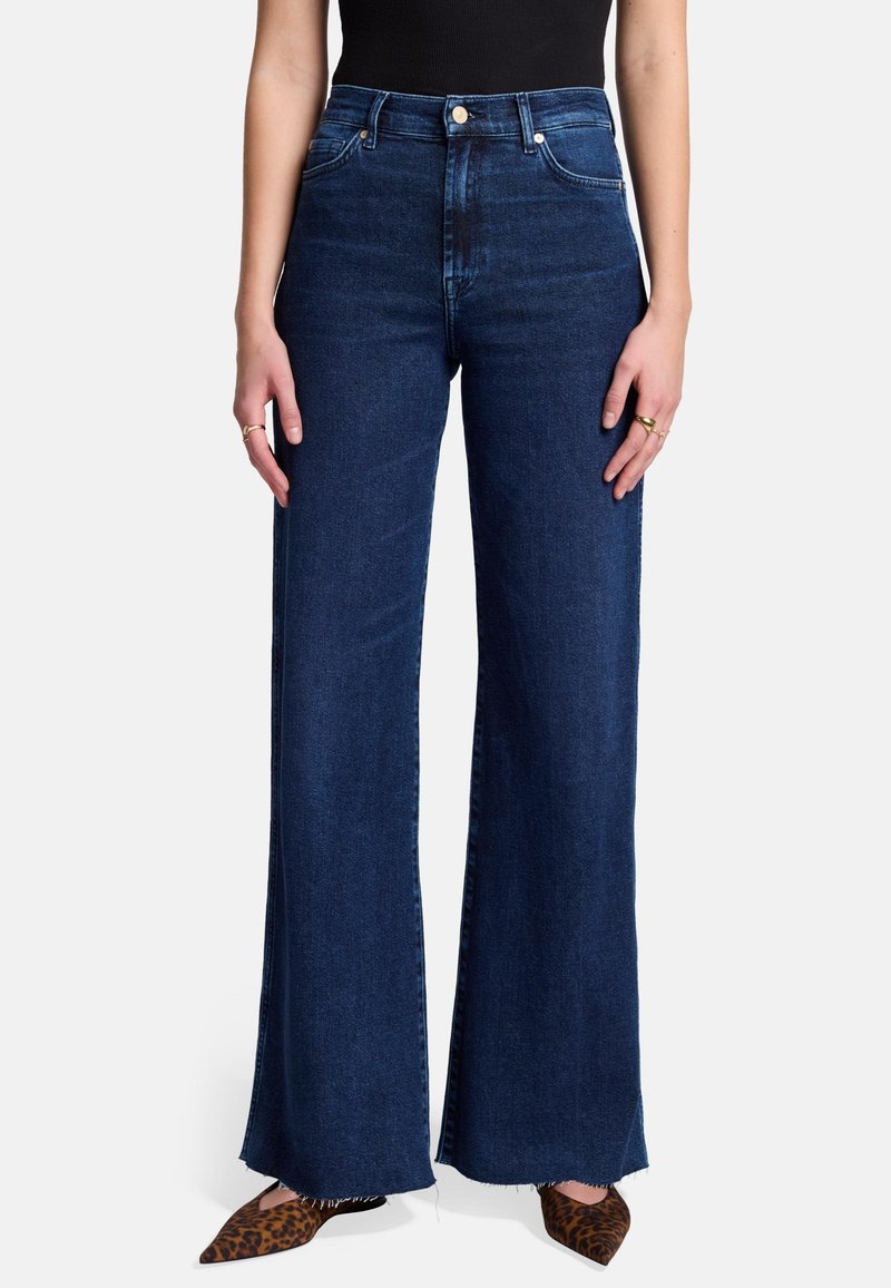 Jeans w wysokim stanie i szerokich nogawkach w ciemnoniebieskim kolorze z surowym wykończeniem, z przednimi kieszeniami i zapięciem na guzik. Połączone z butami w kropki z wężowym nadrukiem.