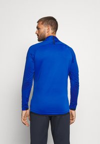 Langsleeve sportshirt in felblauw met een hoge kraag, gestructureerde mouwen en zwarte accenten bij de hals en de zoom.
