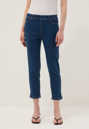 Donna che indossa jeans skinny blu scuro cropped con vita elastica e sandali neri, in piedi davanti a uno sfondo semplice.
