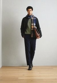 Model nosí tmavú bundu cez zelený sveter, námornícke nohavice a károvaný šál, drží hnedú koženú tašku. Pozadie z drevenej podlahy.