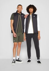 Sorte nylonvester med gule detaljer, grønn skjorte, grå bukser. Uformell fottøy. Modellen poserer sammen i en nøytral bakgrunn.