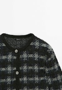 Gros plan d'un cardigan en tricot noir et gris duveteux avec des boutons décoratifs argentés et un col rond sur un fond blanc uni.