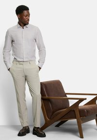 Leicht gestreiftes Button-Down-Hemd aus weichem Stoff, kombiniert mit beigefarbenen maßgeschneiderten Hosen. Braune Lederschuhe vervollständigen den Look. Ein dunkler Holzstuhl ist sichtbar.