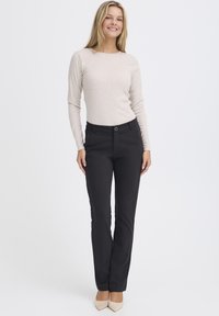 Světlý béžový top s dlouhým rukávem a strukturovaným vzorem, doplněný elegantními černými kalhotami s přední kapsou a poutky na opasek, zvýrazněný neutrálními podpatky.