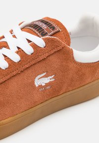 Oranžové semišové tenisky s bílými tkaničkami a akcenty na patě. Obsahují bílé logo Lacoste a značkovou etiketu na jazyku. Gumová podrážka.