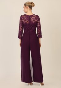 Tuta in pizzo e chiffon di un profondo viola, con maniche a tre quarti, corpino aderente e pantaloni a gamba larga con zip sul retro.