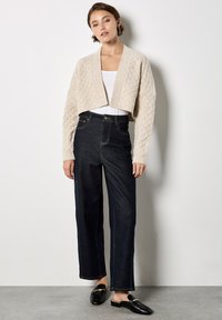 Cardigan corto de punto beige claro sobre una camiseta blanca, combinado con jeans de mezclilla oscuros de talle alto y zapatos negros sin cordones.