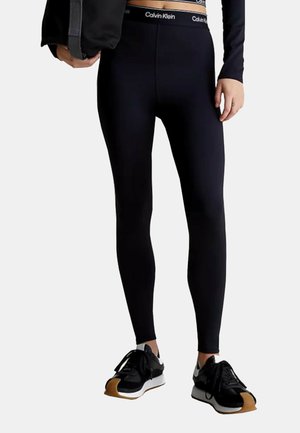 Leggings neri con fascia alta che presenta il logo di Calvin Klein. Texture liscia, design attillato, abbinati a sneakers nere e a una giacca.