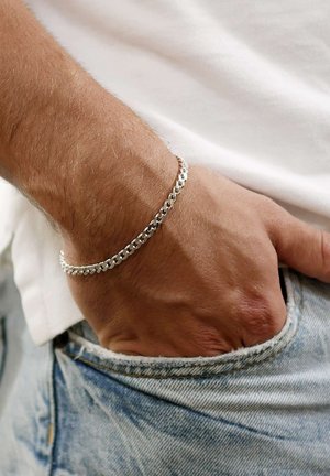 Silver chain bracelet on a wrist, featuring interlocking links with a polished finish. Worn on a hand resting in a jean pocket.