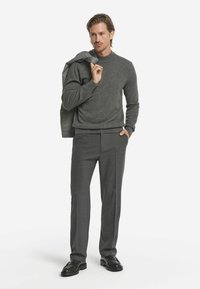 Grå ulltröja med rund halsringning, matchad med skräddarsydda grå byxor och svarta loafers, kompletterad med en grå kavaj som hänger över ena axeln.