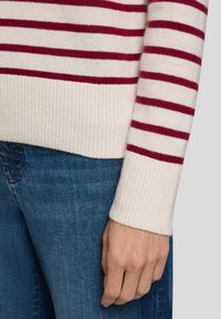 Cremiger Pullover mit weinroten horizontalen Streifen und gerippten Bündchen, gezeigt an einer Hand. Kombiniert mit blauem Denim, um Textur und Farbkontrast hervorzuheben.