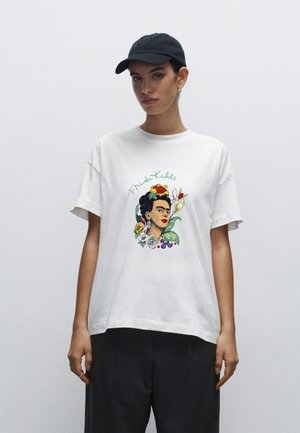 Hvid bomulds-T-shirt med en farverig Frida Kahlo-illustration med blomsterdetaljer og grøn tekst. Modellen bærer en sort kasket og mørke bukser.