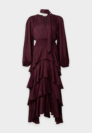 Donna che indossa un lungo abito bordeaux a strati con maniche lunghe e scollo annodato, abbinato a sandali neri con tacco e punta aperta.