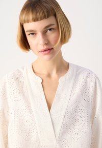 Blusa bianca in tessuto leggero con intricati motivi a taglio, vestibilità morbida e scollatura a V con bordi ondulati. Texture sottile in tutto il capo.